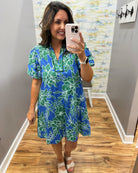 Woman taking a mirror selfie wearing a blue floral dress in a room with wooden flooring and a colorful wall.
