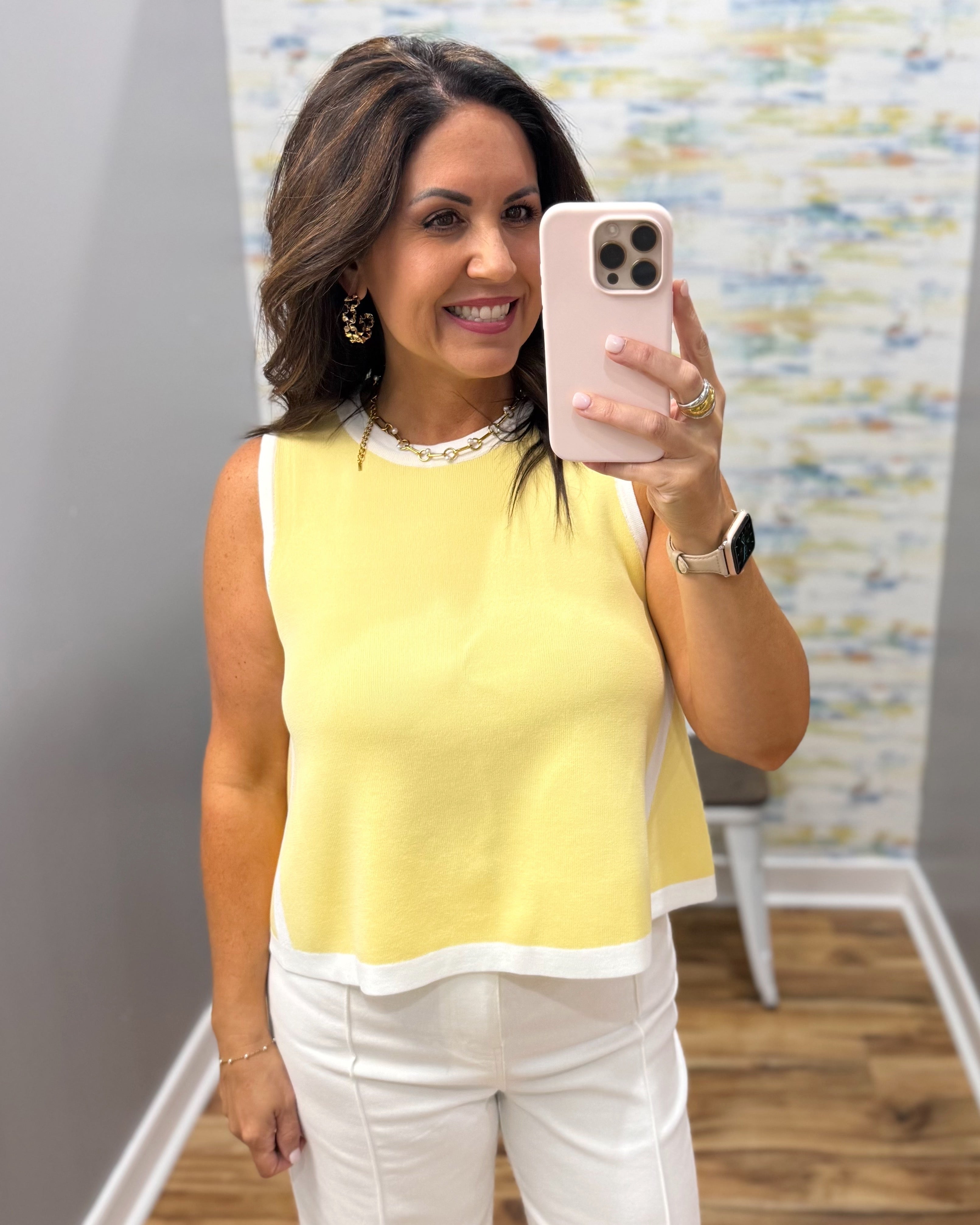 Woman taking a selfie in a yellow top and white pants, holding a phone.