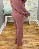 A person wearing a dusty rose pink two-piece lounge set consisting of a long-sleeve top and matching wide-leg pants, standing on a wood floor in front of a white bench.