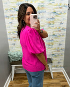 Woman taking a mirror selfie wearing a bright pink top and blue jeans in front of a textured wall.