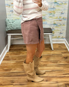 A person wearing a chestnut color wrap-style mini skirt with a single button closure, paired with a pink and white striped long-sleeve top and Corkys City Slicker boots in Sand Faux Suede.