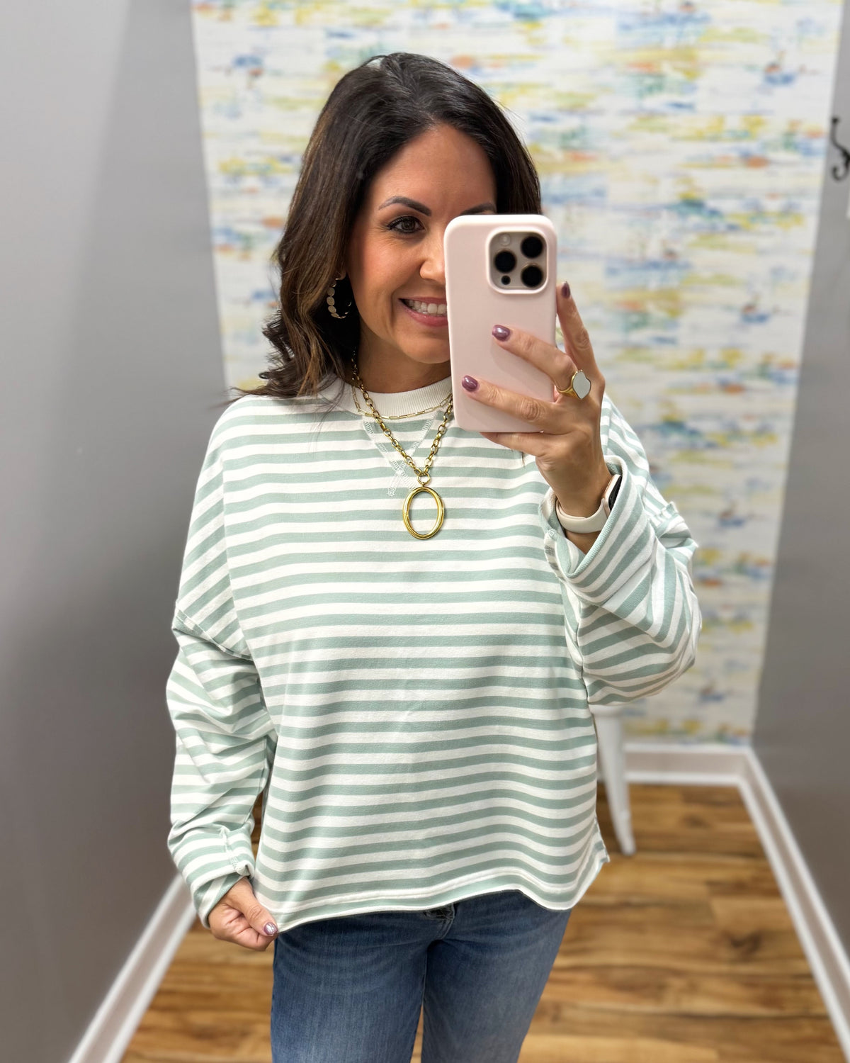 A woman taking a mirror selfie wearing a light green and white horizontally striped long-sleeve top, blue jeans, and gold jewelry.