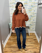 A woman takes a mirror selfie in a fitting room wearing a rust-brown long-sleeve knit sweater with a scalloped white hem, dark blue jeans, and tan boots.