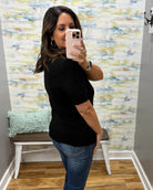 A woman taking a mirror selfie wearing a black short-sleeve crewneck top and blue jeans, standing in a room with a patterned wallpaper background.
