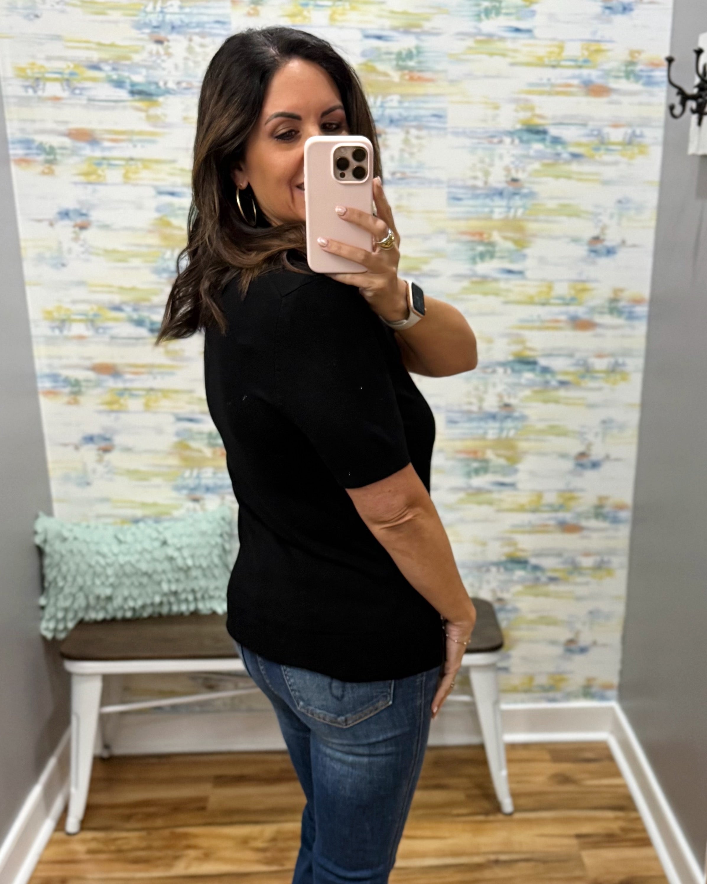A woman taking a mirror selfie wearing a black short-sleeve crewneck top and blue jeans, standing in a room with a patterned wallpaper background.