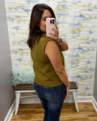 A woman takes a mirror selfie in a fitting room wearing an olive green sleeveless ribbed knit sweater vest and blue jeans.