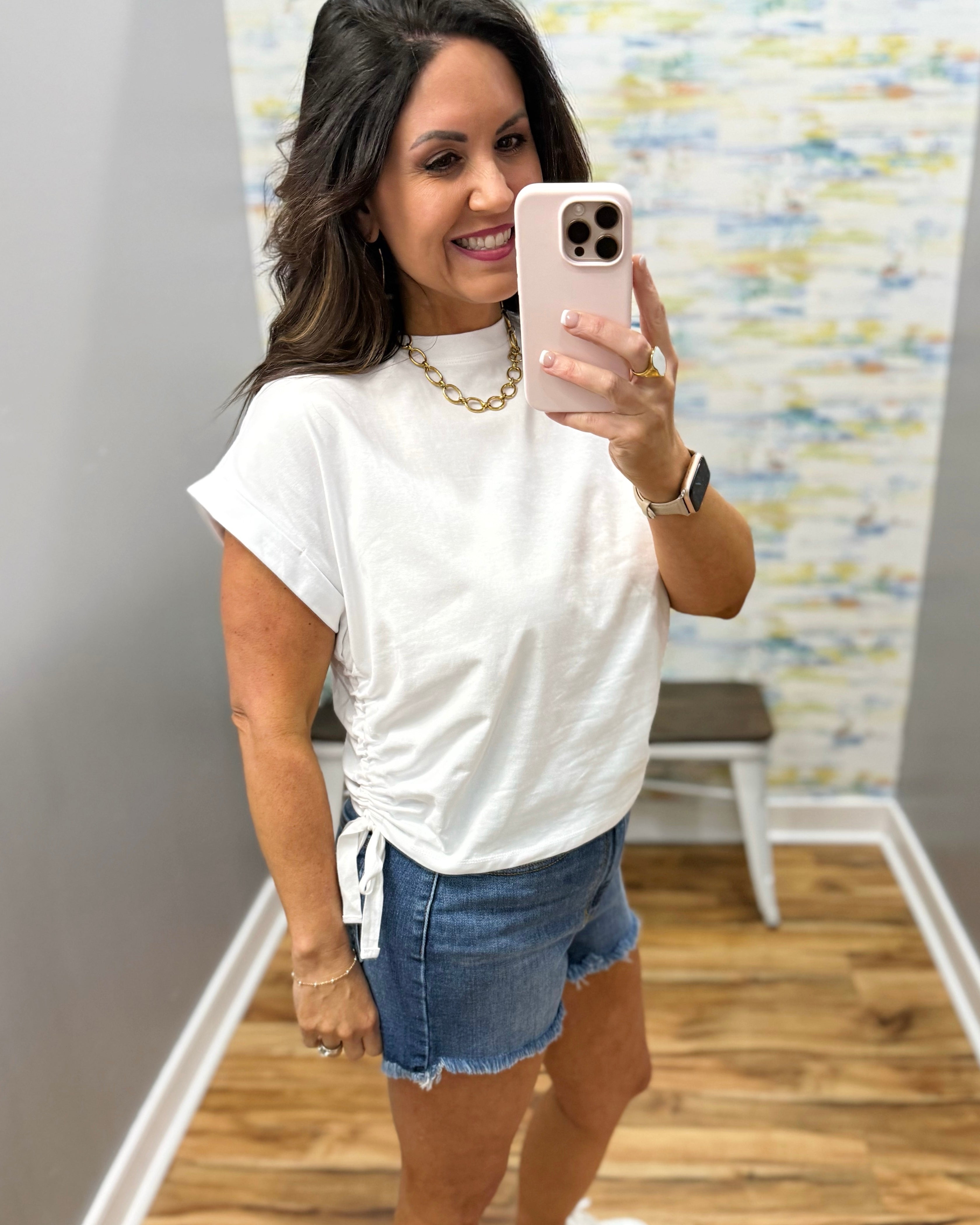 Woman taking a mirror selfie wearing a white t-shirt and denim shorts in a room with a colorful wall.