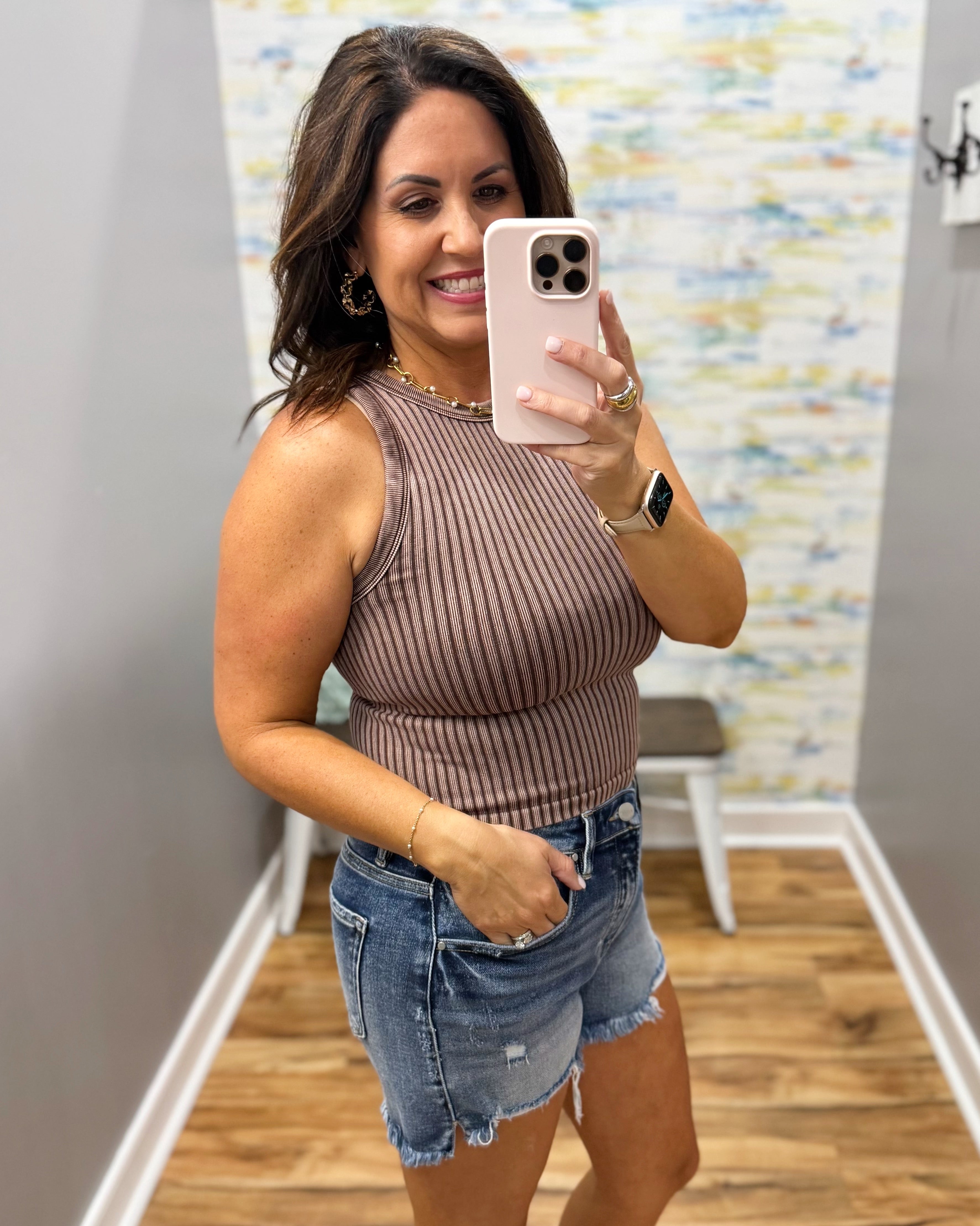 Woman taking a mirror selfie wearing a ribbed tank top and denim shorts in a room with a colorful wall.