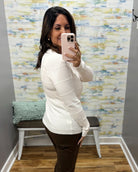 A woman takes a mirror selfie in a fitting room wearing a white ribbed knit long-sleeve sweater and dark brown pants.