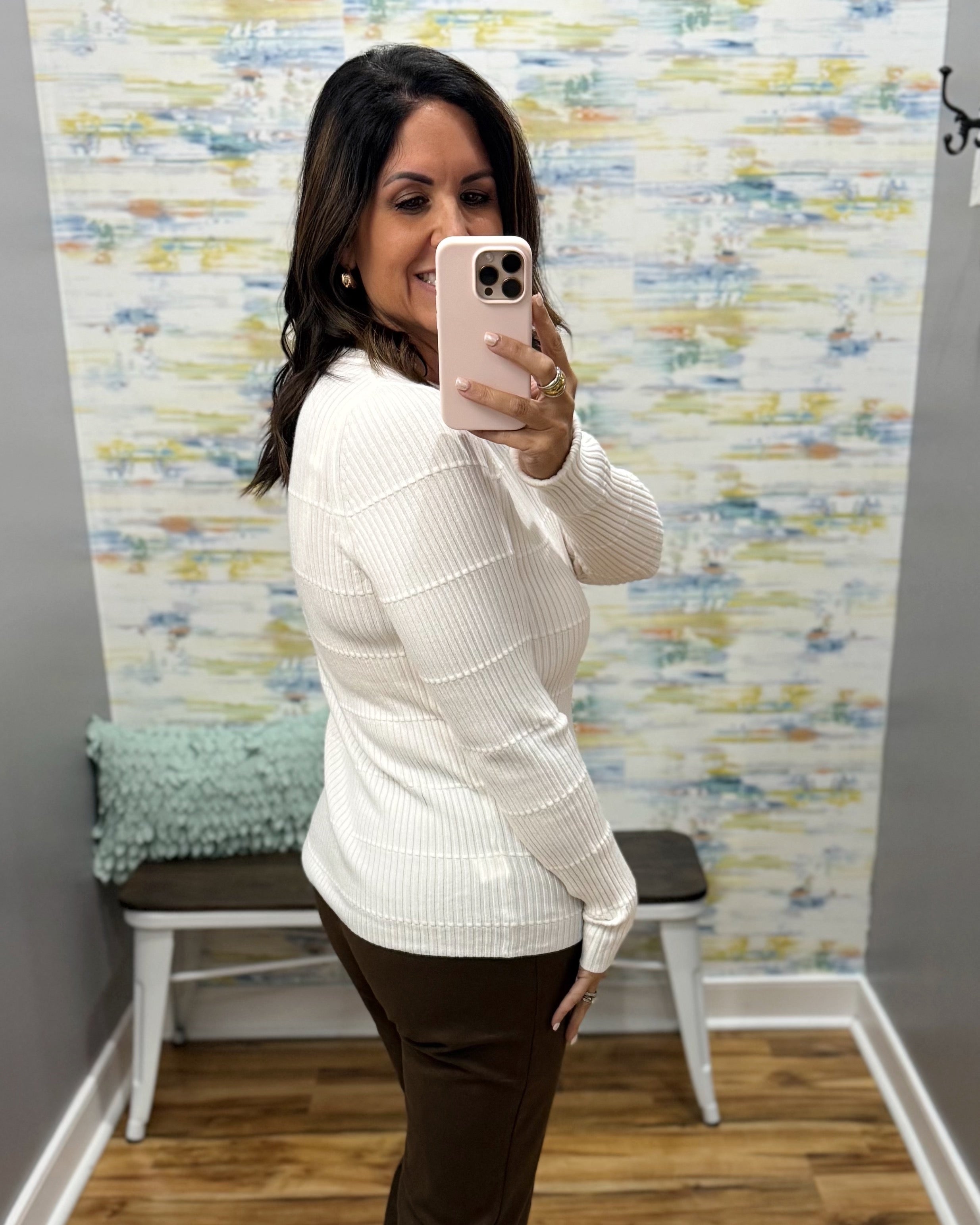 A woman takes a mirror selfie in a fitting room wearing a white ribbed knit long-sleeve sweater and dark brown pants.