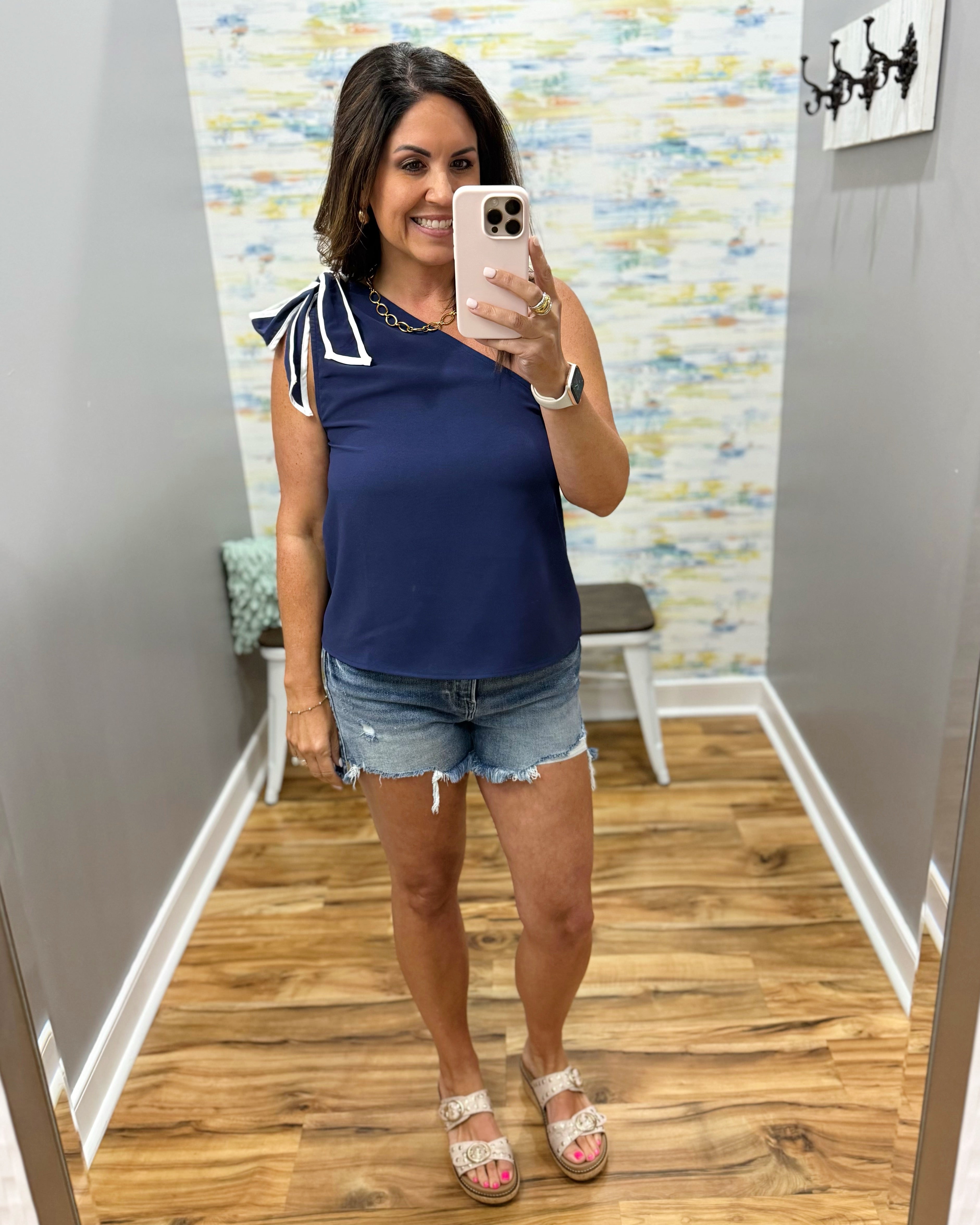 Woman taking a mirror selfie wearing a navy top with bow sleeves and denim shorts in a room with a colorful wall.