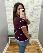 A woman takes a mirror selfie in a fitting room wearing a short-sleeve knit top in plum with light pink polka dots and pink trim, paired with blue jeans.