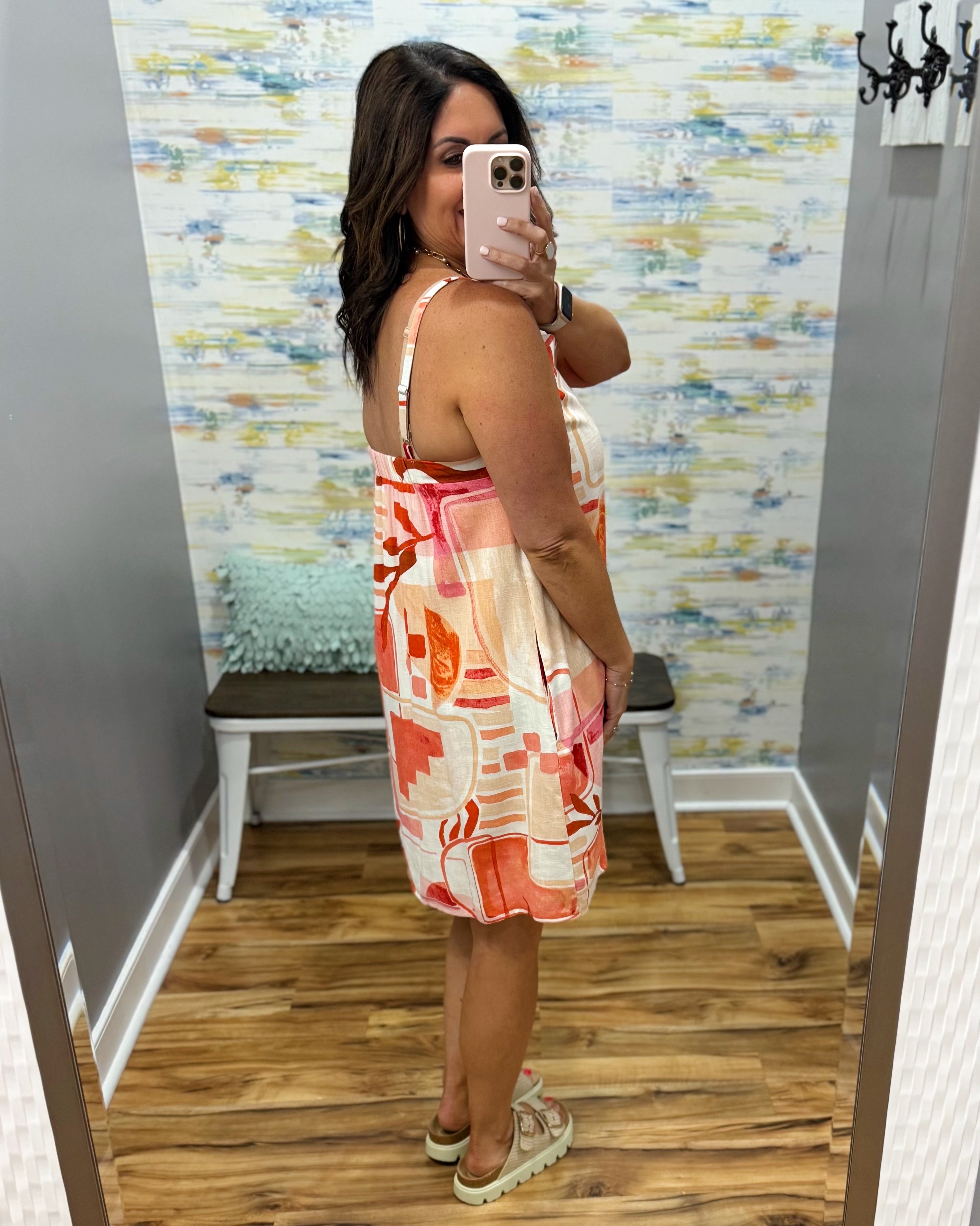 Woman taking a mirror selfie wearing a colorful dress in a room with patterned wallpaper.