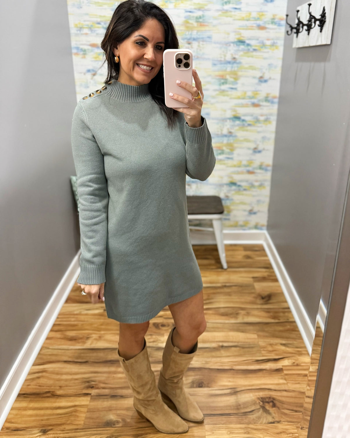 A woman takes a mirror selfie wearing a sage green mock-neck knit sweater dress with gold button details on the shoulders, paired with beige suede boots, in a home setting with a wood floor and abstract wall art.