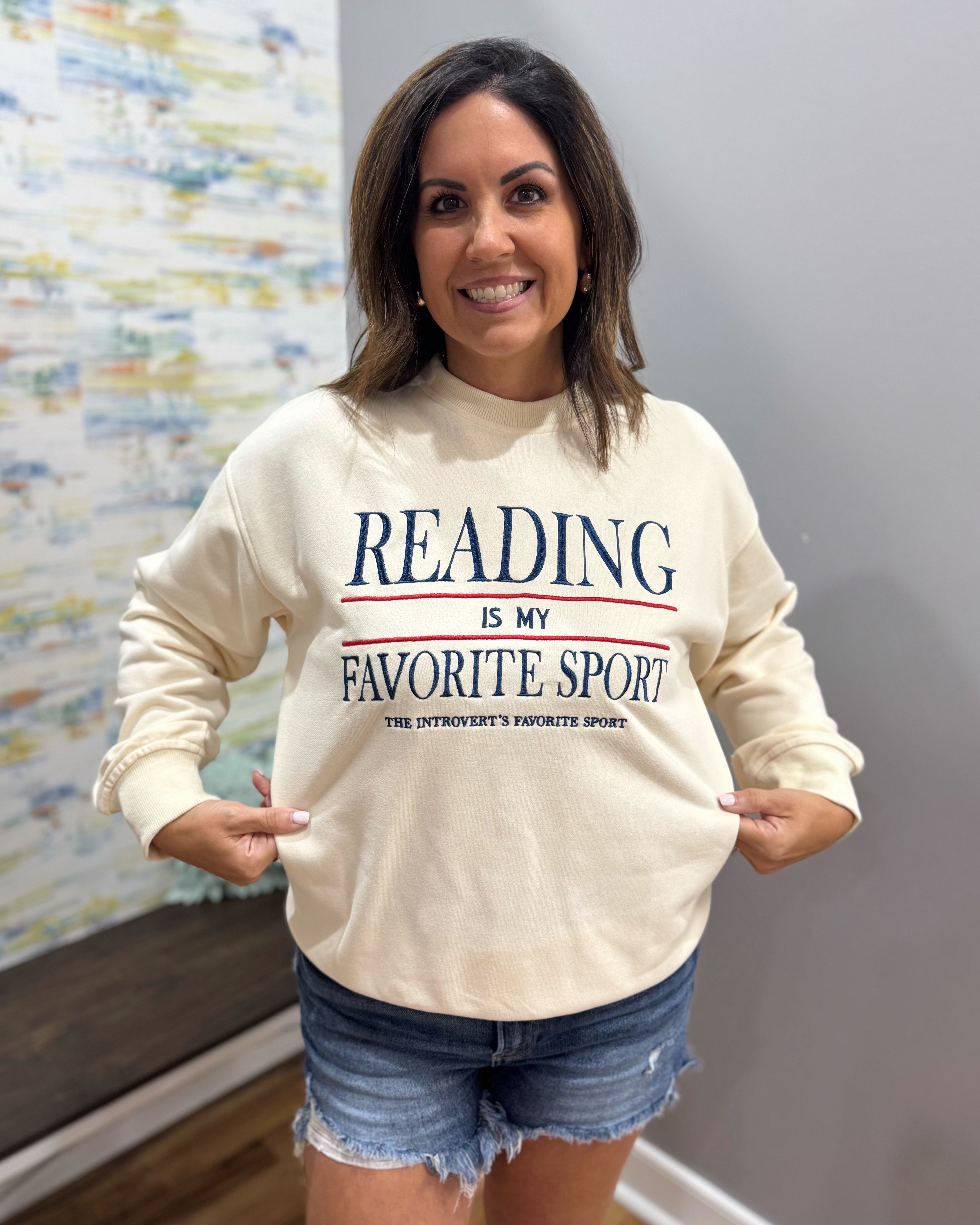 A woman wearing a cream-colored crewneck sweatshirt with the text 'READING IS MY FAVORITE SPORT' in navy blue and red, paired with denim shorts, standing in an indoor setting.