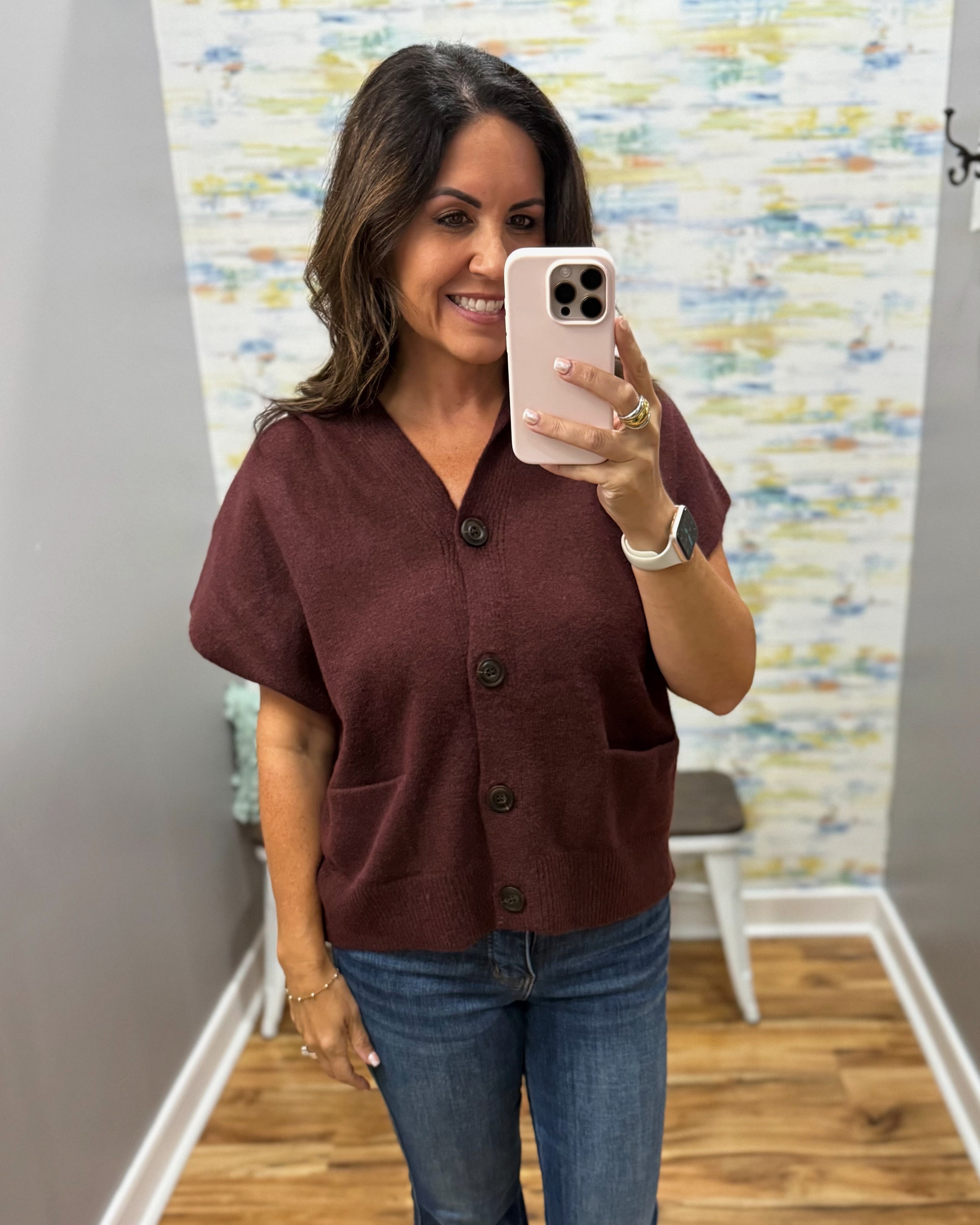 A woman taking a mirror selfie in a boutique wearing a short-sleeve, v-neck knit top in a deep burgundy color with dark buttons and front pockets, paired with blue jeans.