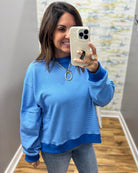 A woman takes a mirror selfie wearing a light blue and royal blue horizontally striped long-sleeve sweatshirt, paired with gray jeans and a gold chain necklace.