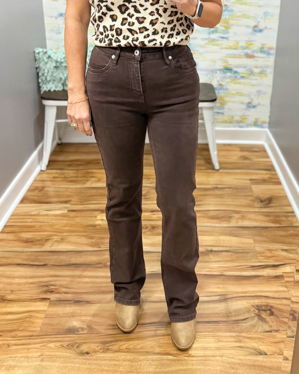 A person wearing mica dark brown straight-leg jeans, a leopard-print top, and metallic gold boots, standing on a wood floor in an indoor room.