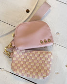 A small pink pebbled leather pouch with gold-tone hardware and a matching pink woven strap, placed on a white distressed wood surface alongside a pink-and-beige checkered zip pouch.