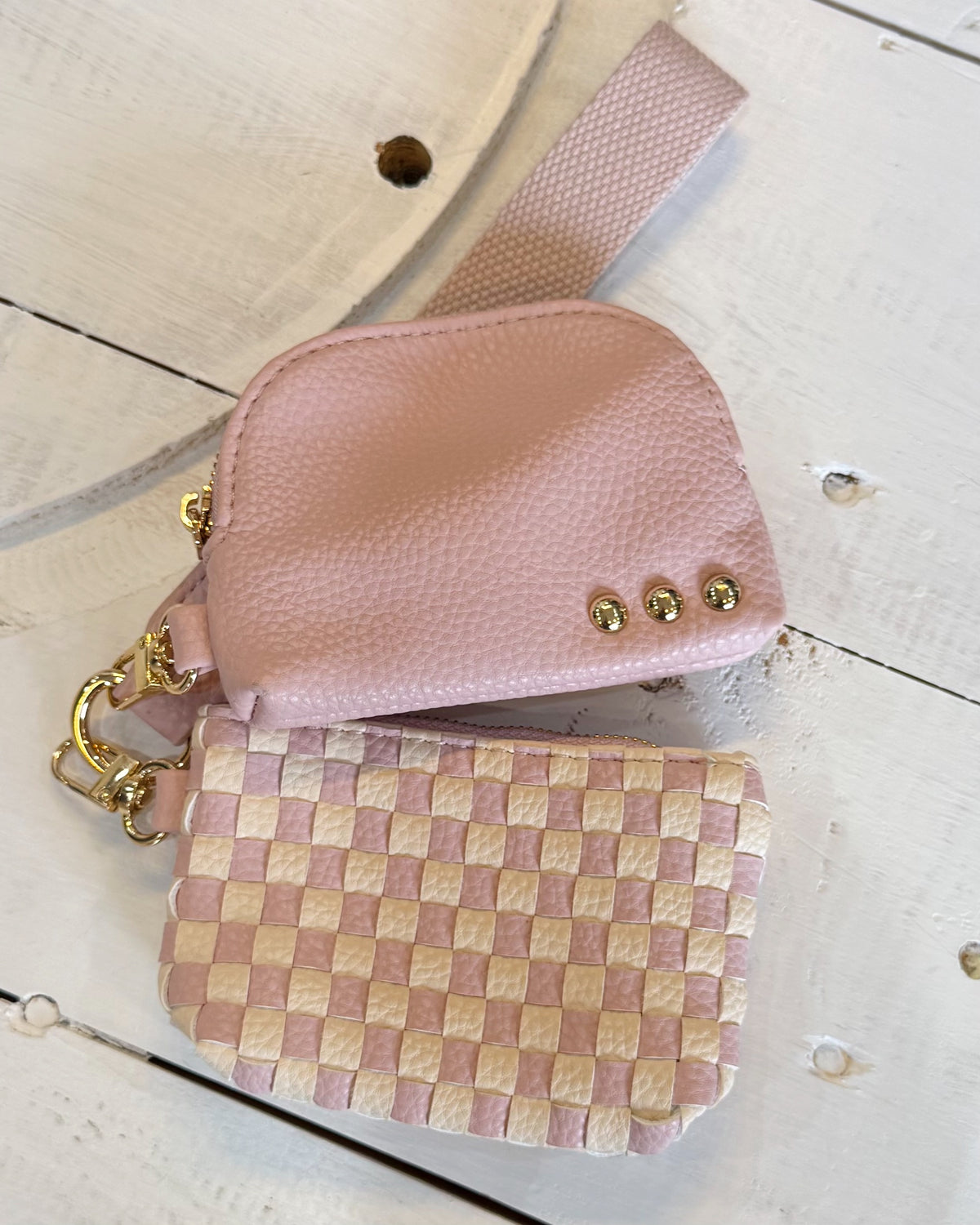 A small pink pebbled leather pouch with gold-tone hardware and a matching pink woven strap, placed on a white distressed wood surface alongside a pink-and-beige checkered zip pouch.