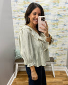A woman takes a mirror selfie wearing a light sage green and white vertically striped blouse with ruffled details on the collar and cuffs, paired with dark blue jeans.