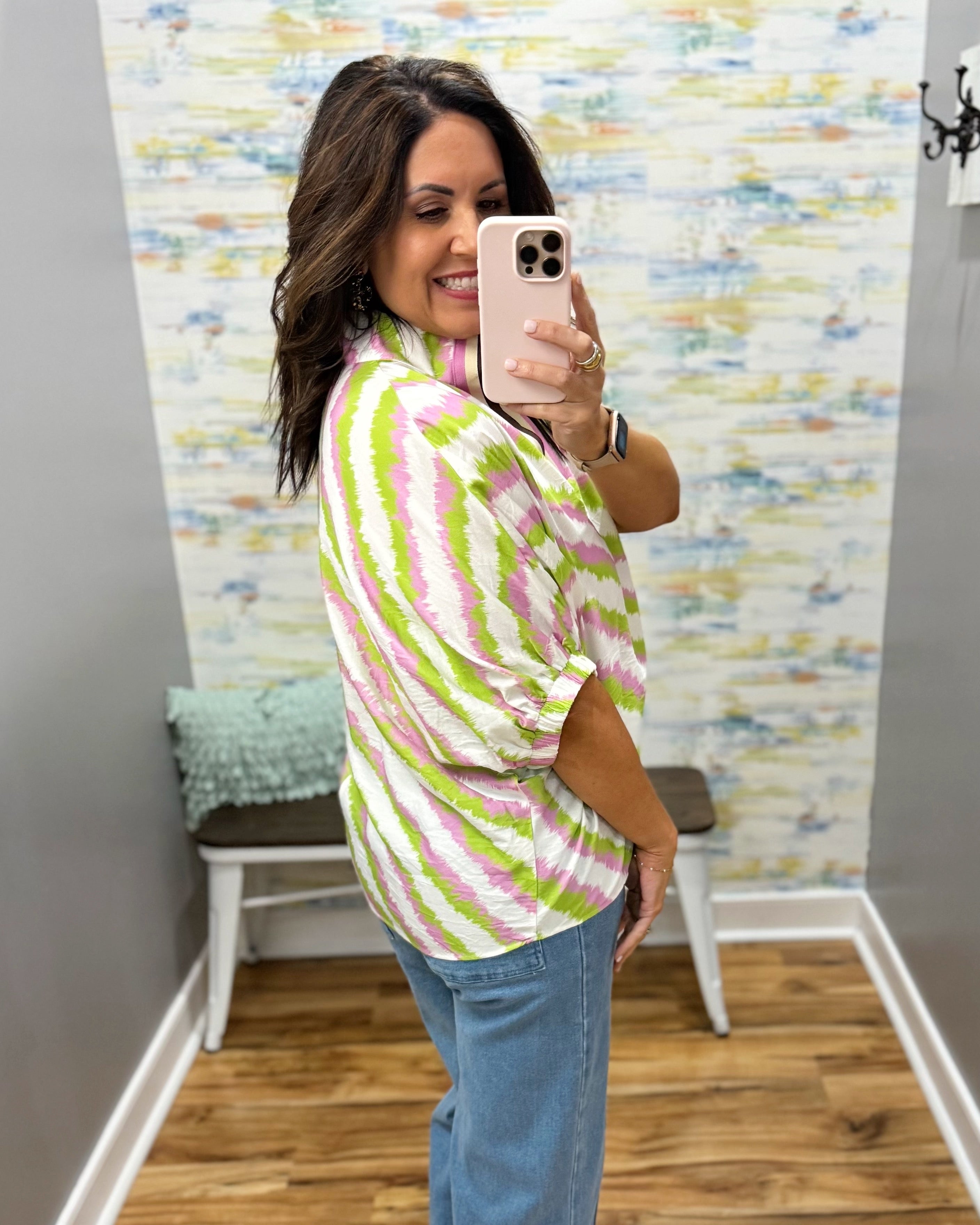 Woman taking a mirror selfie wearing a colorful striped shirt and blue jeans in a room with patterned wallpaper.