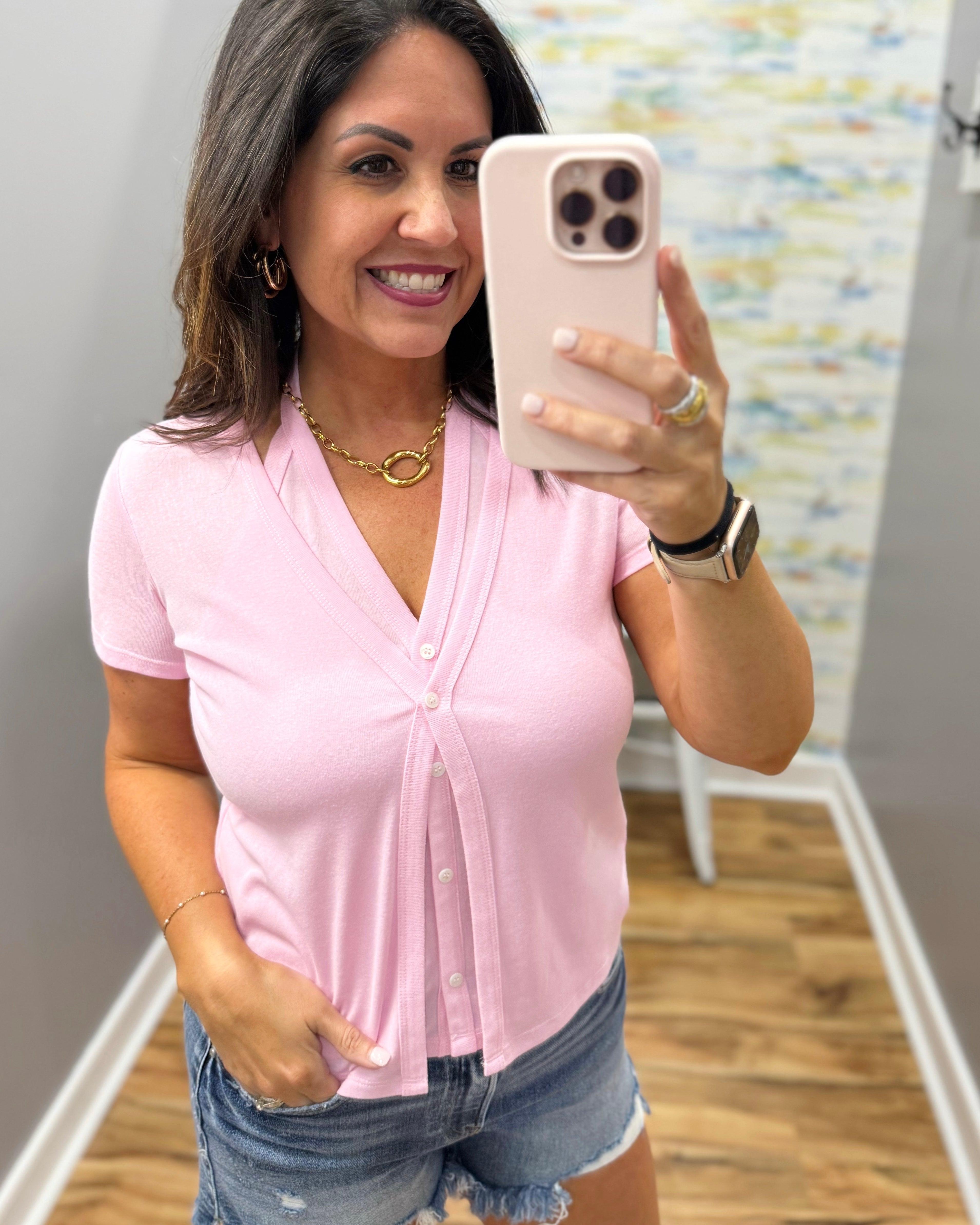 Woman taking a mirror selfie wearing a pink top and denim shorts.