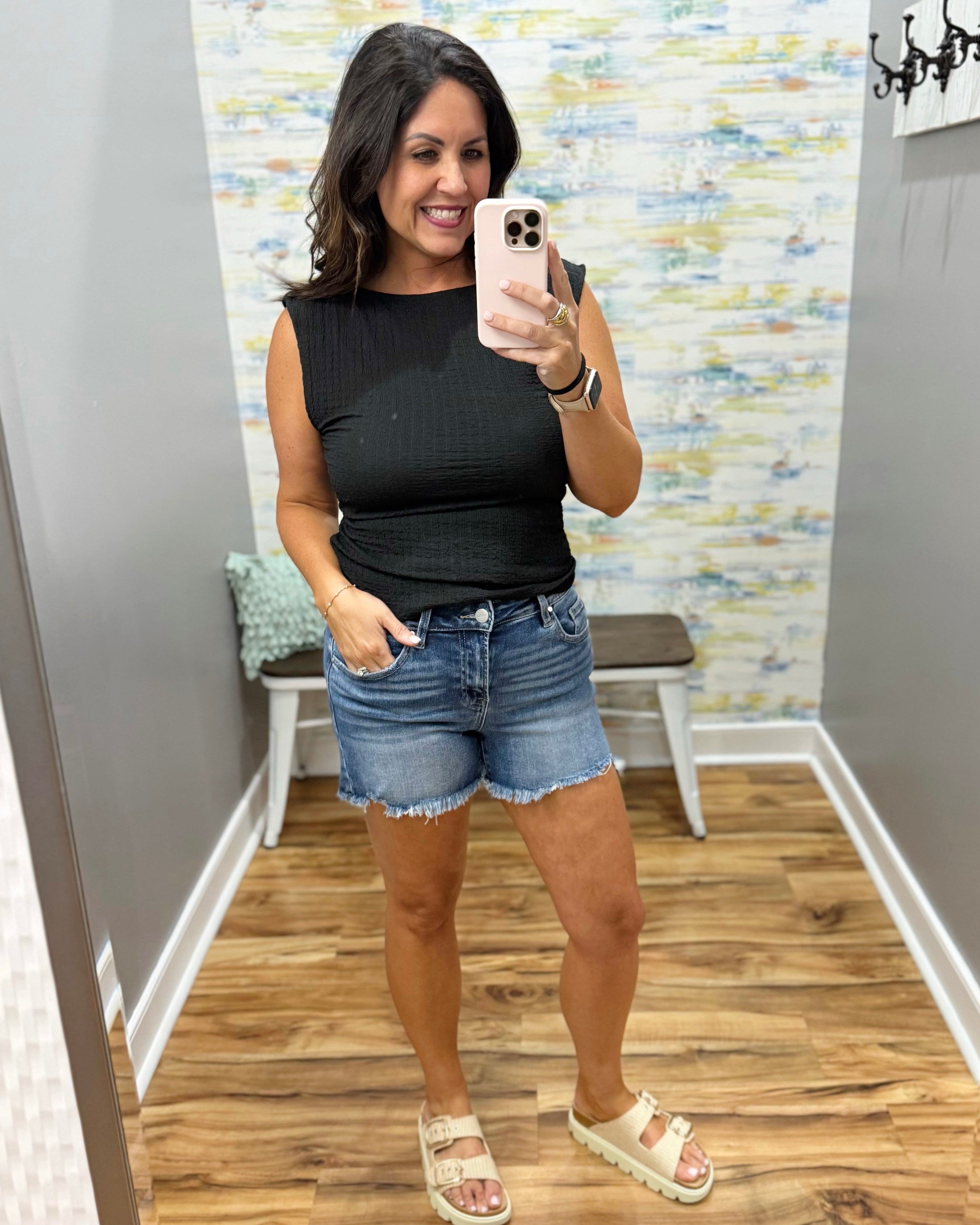 Woman taking a mirror selfie wearing a black sleeveless top and denim shorts in a room with a patterned wall.