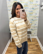 A woman takes a mirror selfie wearing a beige and white striped knit sweater, blue jeans, and a light pink phone case in a fitting room with wood floors and patterned wallpaper.