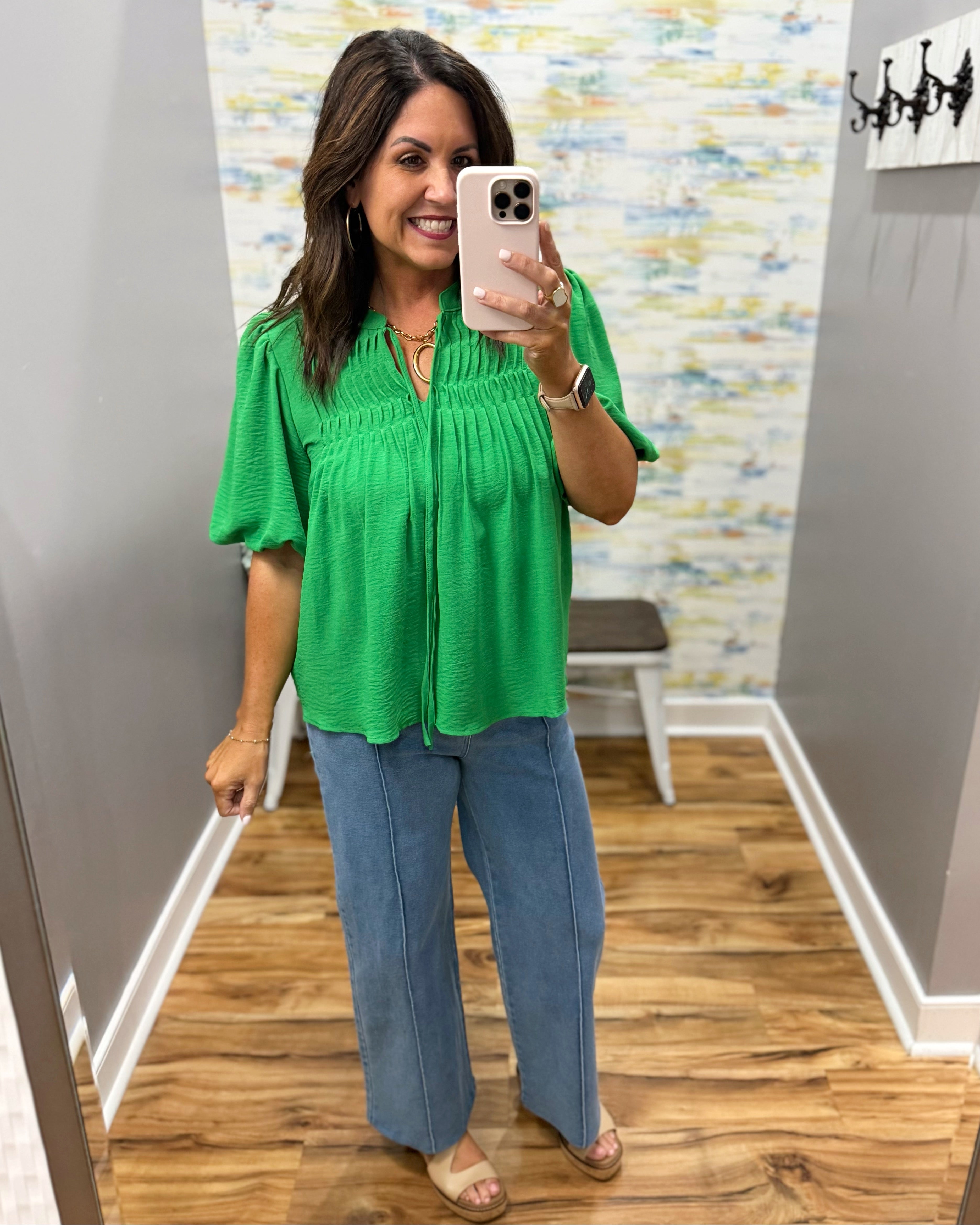 Woman taking a mirror selfie wearing a green top and blue jeans in a store.