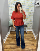 A woman takes a mirror selfie in a fitting room wearing a rust-orange short-sleeve peplum top and dark blue flare jeans.