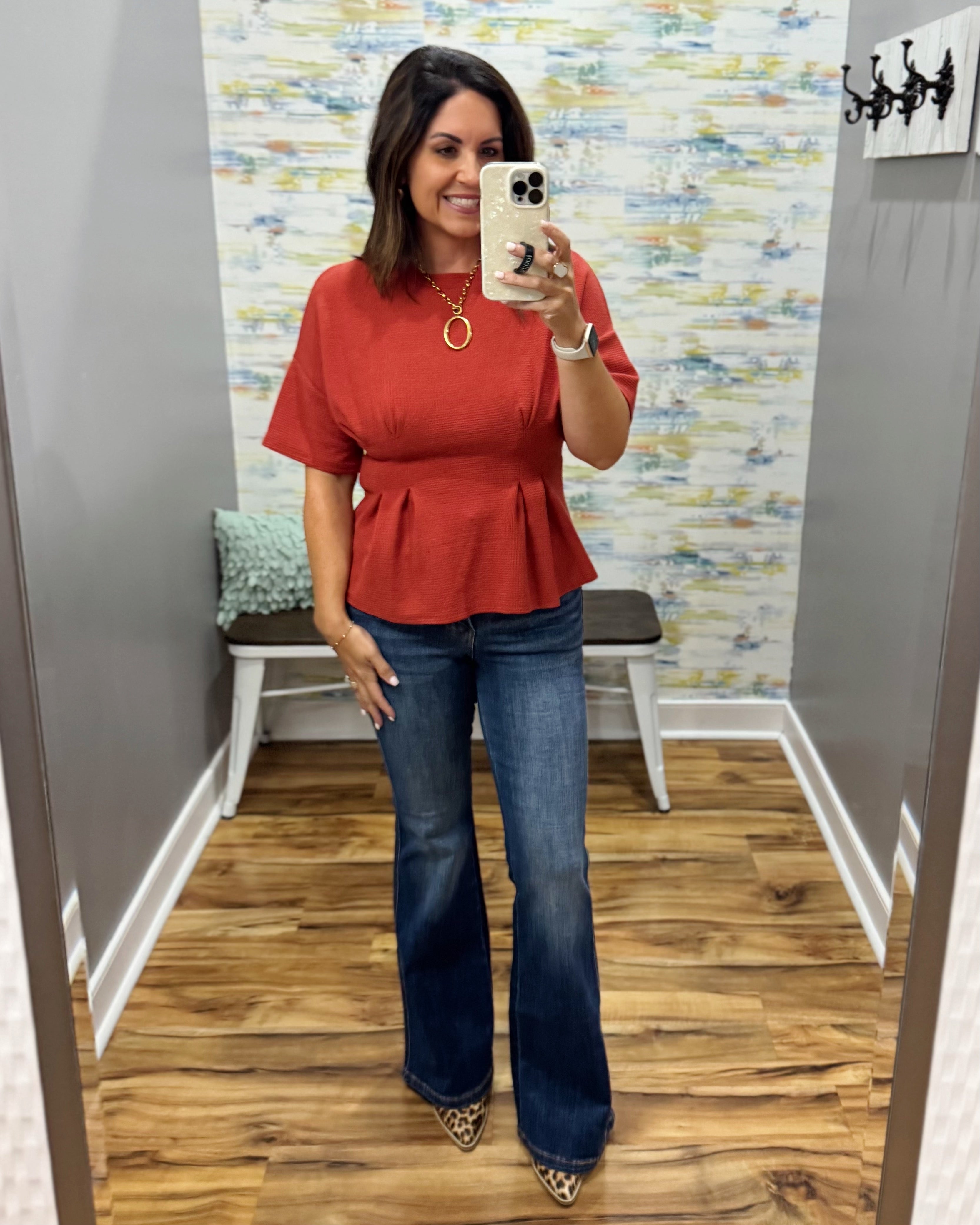 A woman takes a mirror selfie in a fitting room wearing a rust-orange short-sleeve peplum top and dark blue flare jeans.