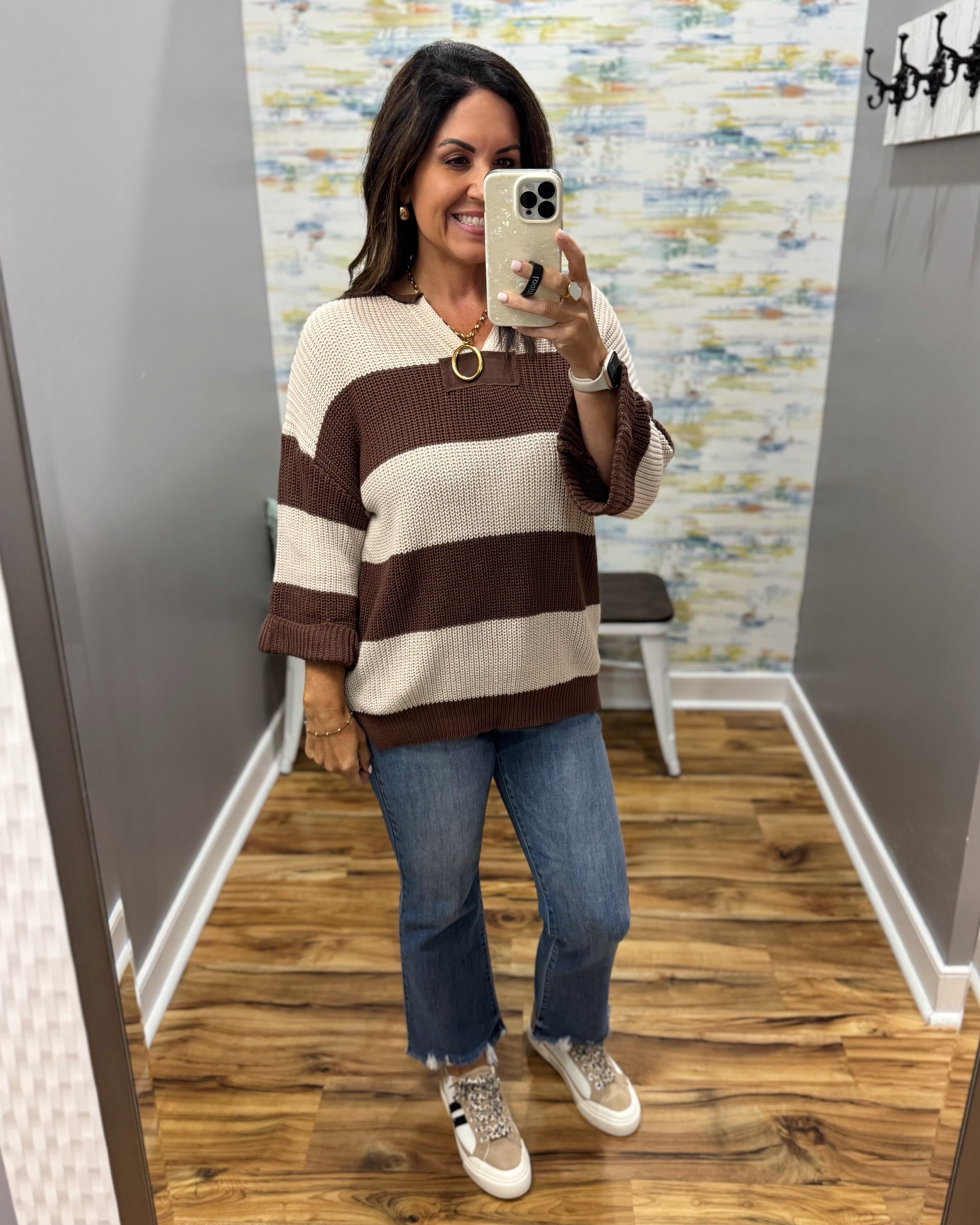 A woman takes a mirror selfie in a fitting room wearing a brown and cream striped knit sweater, blue jeans, and beige sneakers.
