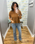 A woman takes a mirror selfie wearing a brown short-sleeve textured top with ruffle details, paired with light-wash blue jeans and tan boots, in a fitting room.