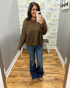 A woman takes a mirror selfie wearing a brown knit crewneck sweater with a chest pocket, blue flare jeans, and beige pointed-toe flats in a fitting room with wood floors and a patterned wall.