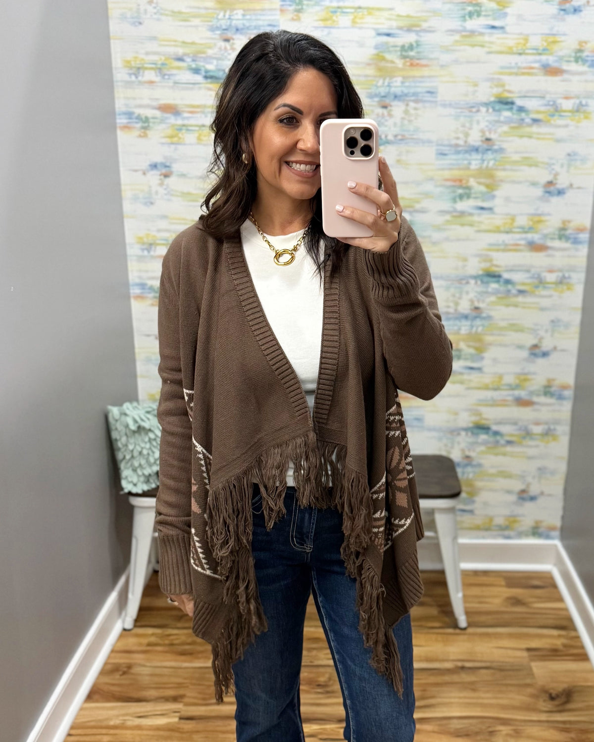 A woman takes a mirror selfie in a fitting room wearing a brown open-front knit cardigan with fringe and decorative knit details, a white top, and blue jeans.