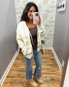 A woman takes a mirror selfie in a fitting room wearing a cream and tan patterned knit zip-up hoodie over a brown top, blue jeans, and beige sneakers.