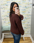 A woman takes a mirror selfie wearing a dark brown ribbed knit sweater and blue jeans in front of a patterned wall.