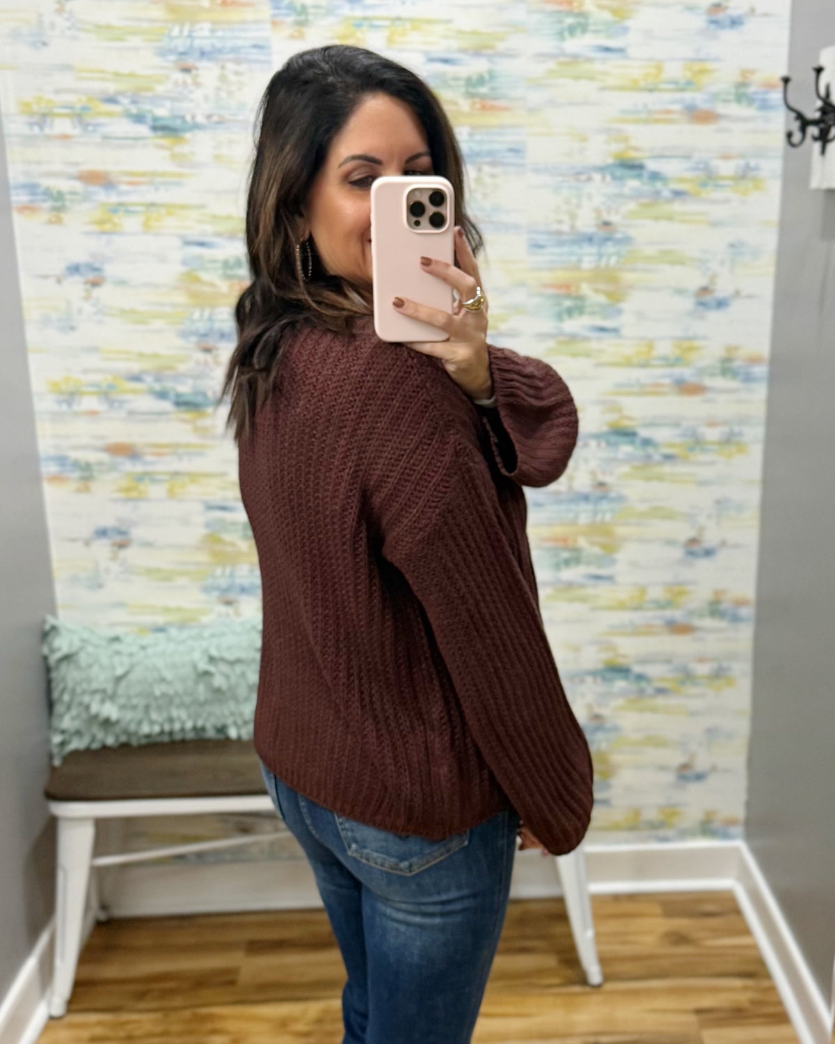 A woman takes a mirror selfie wearing a dark brown ribbed knit sweater and blue jeans in front of a patterned wall.