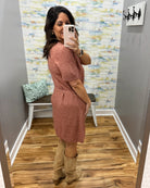 A woman takes a mirror selfie in a fitting room wearing a dusty rose short-sleeve knit dress with a tie waist and tan knee-high boots.