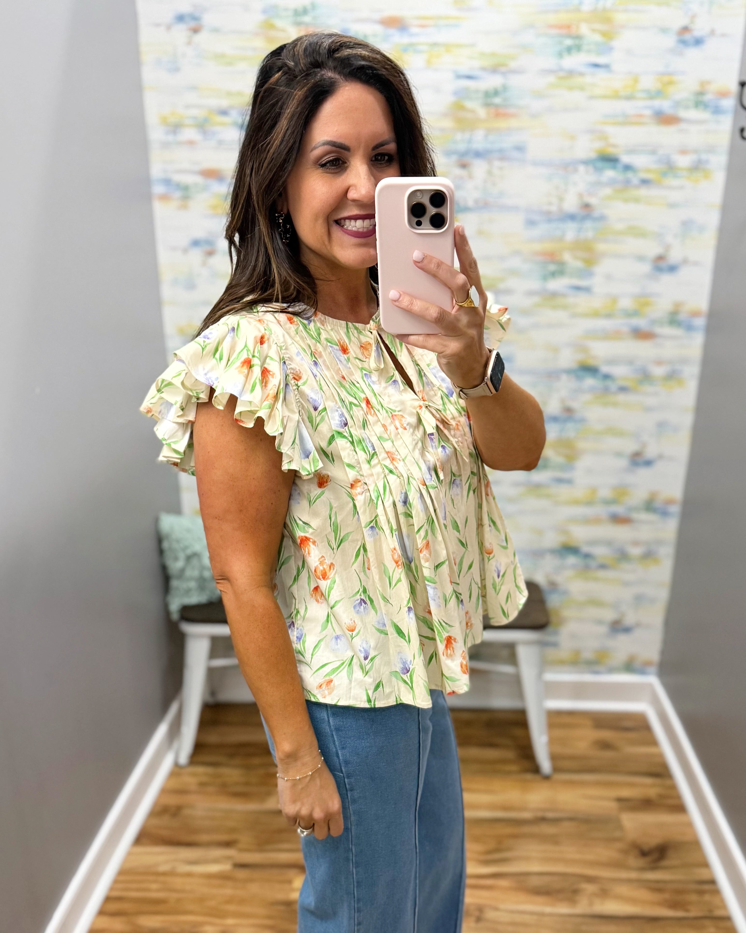 Woman taking a mirror selfie wearing a floral blouse and blue jeans in a room with a colorful wall.