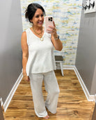 Woman taking a mirror selfie wearing a white top and light gray pants in a room with a patterned wall.
