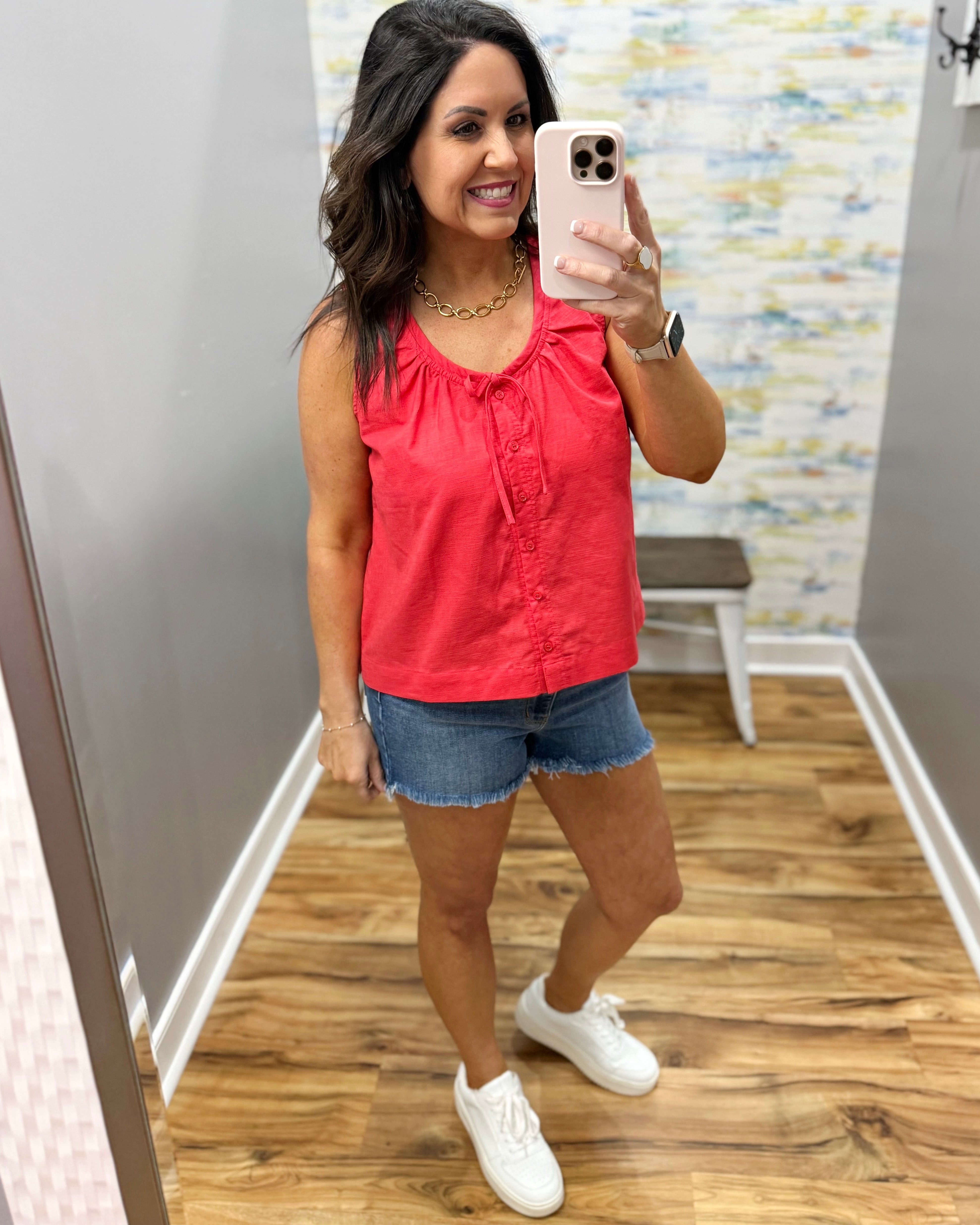 Woman taking a mirror selfie wearing a red tank top and denim shorts.