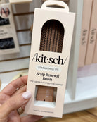 A hand holding a Kit Schi scalp renewal brush package, showing the retail box with a clear window revealing the brown nylon brush inside.