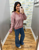 A woman takes a mirror selfie in a fitting room wearing a pink ribbed knit sweater with a hood and drawstrings, paired with blue jeans and beige shoes.