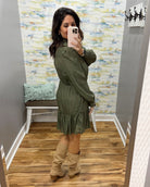 A woman takes a mirror selfie in a fitting room wearing an olive green long-sleeve mini dress with a ruffled hem and tan knee-high boots.