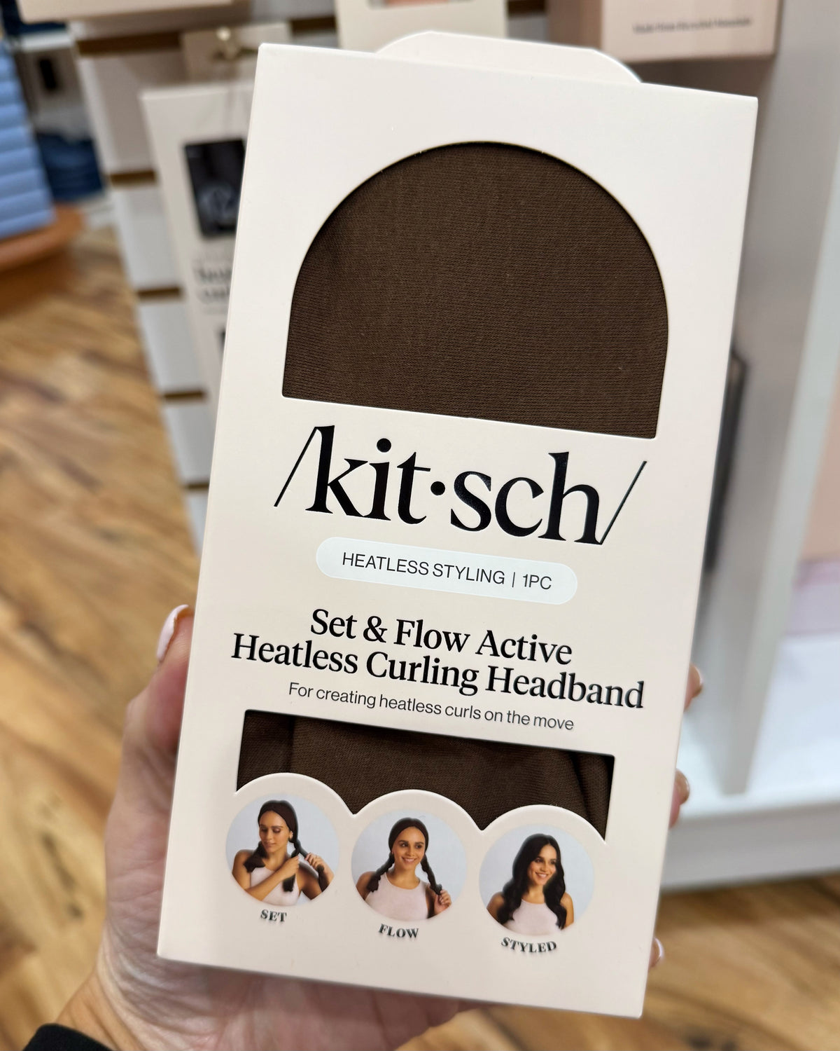 A hand holding a white retail box for a Kit-Sch heatless curling headband. The box has a large arched window showing the dark brown headband inside and text reading 'Kit-Sch' and 'Set & Flow Active Heatless Curling Headband.
