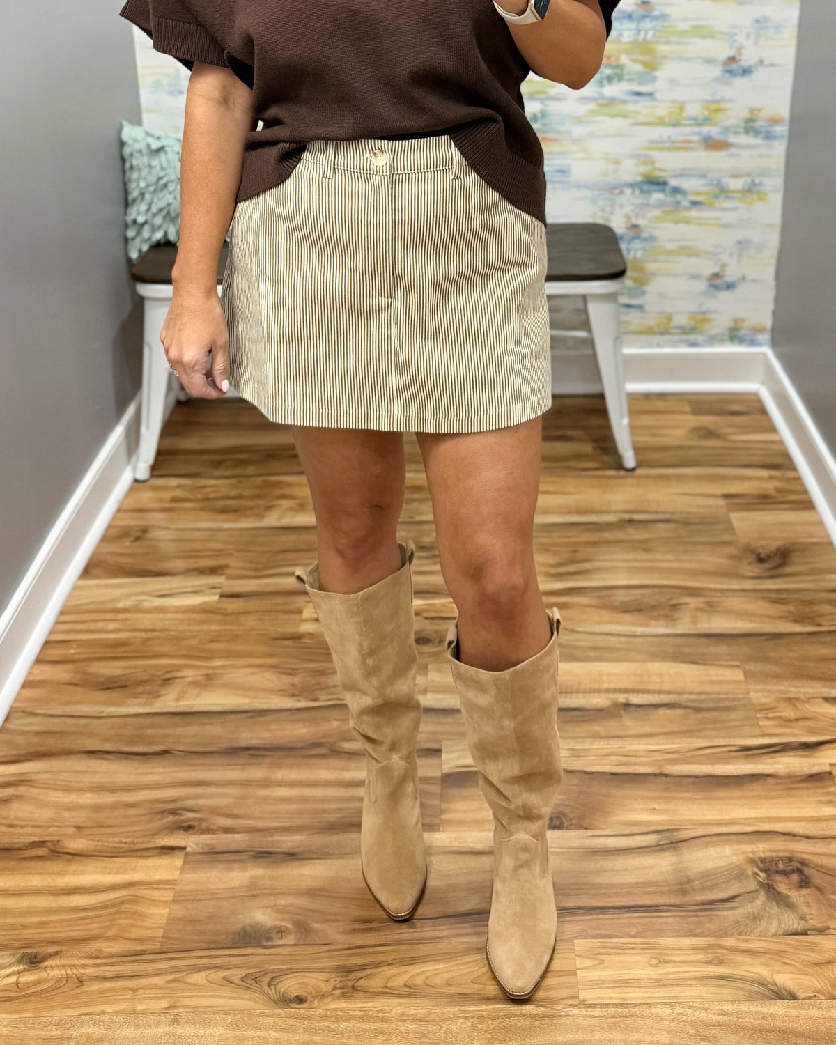 A person wearing a beige corduroy mini skirt, brown top, and tan knee-high suede boots, standing on a wood floor in an indoor room.