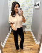 A woman takes a mirror selfie in a fitting room wearing a beige short-sleeve knit top and black pants with leopard print flats.
