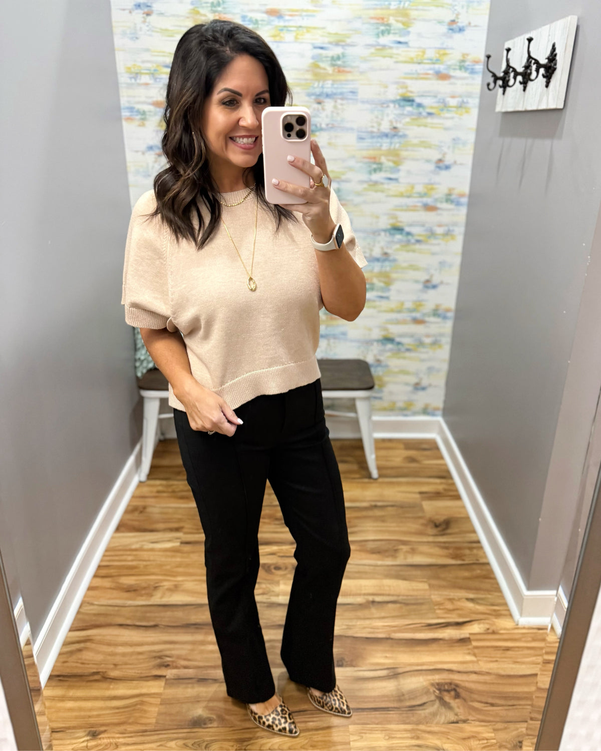 A woman takes a mirror selfie in a fitting room wearing a beige short-sleeve knit top and black pants with leopard print flats.